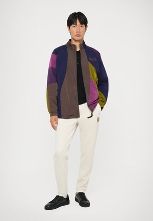 Colorful zip-up jacket featuring purple, brown, and green panels; paired with cream sweatpants and black sneakers. Smooth texture and casual fit.