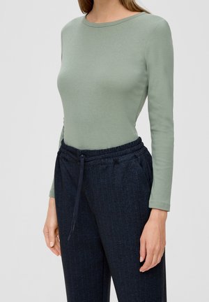 Femme portant un haut ajusté manches longues vert sauge et un pantalon taille haute bleu foncé à cordon, sur un fond uni.