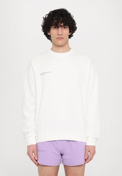 Hvid sweatshirt med rund hals og lange ærmer, prydet med en lille trykt detalje på brystet. Sammen med lavendelshorts.