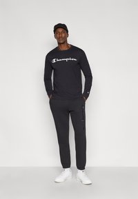 Μαύρο μακρυμάνικο πουκάμισο με λευκό λογότυπο "Champion", συνδυασμένο με σκούρο γκρι φόρμες με λογότυπο στο πλάι, φορεμένο με λευκά αθλητικά παπούτσια.