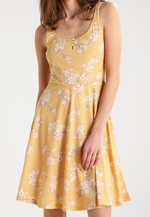 Ärmelloses gelbes Kleid mit rosa-weißem Blumenmuster, Rundhalsausschnitt und drei kleinen Knöpfen an der Brust, getragen von einer Frau.