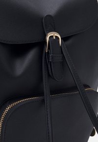 Černý kožený batoh s hladkou texturou, který má zapínání na zlatou sponu, nastavitelné popruhy a přední kapsu na zip.