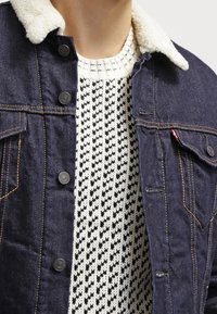 Veste en denim avec un col sherpa doux de couleur crème, dotée de boutons foncés et de coutures dorées, portée sur un pull à motifs crème et noir.