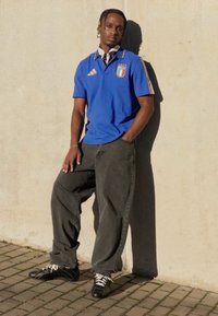 Μπλε μπλουζάκι πόλο με λογότυπο της ιταλικής σημαίας, γκρι φαρδιά παντελόνια και μαύρα αθλητικά παπούτσια, ακουμπισμένος σε ένα ανοιχτόχρωμο τοίχο.