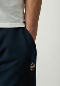 Pantaloni sportivi blu navy con una texture liscia e tasca laterale, dotati di una patch rotonda in bordeaux e grigio etichettata "COLMAN."