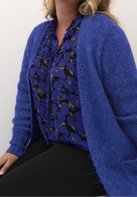 Lila Wolljacke über einer blauen Bluse mit schwarz-blumigem Muster und grünen Akzenten. Model sitzt und trägt schwarze Hosen.
