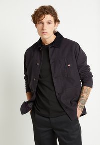 Giacca nera con colletto in tessuto spesso, con chiusura a bottoni frontale, tasca sul petto e foderatura a quadri in contrasto. Il modello indossa una t-shirt nera sotto.