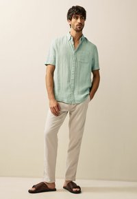 Light green short-sleeved button-up shirt with a chest pocket, paired with light beige pants and brown slide sandals on a neutral backdrop.