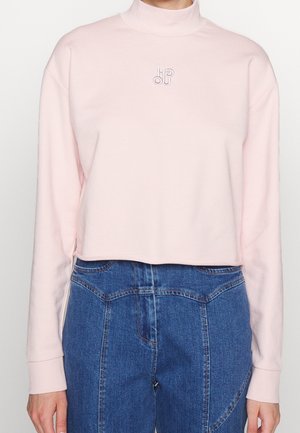 Pull court à col roulé rose clair à manches longues avec petit logo brodé, associé à un jean bleu taille haute.