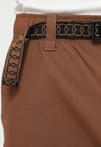 Hnedé nohavice so zberaným pásom, ktoré majú textúrovaný čierno-hnedý opasok a plochú pracku. Žiadne viditeľné vrecká.