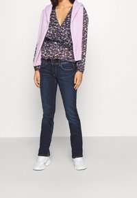 Blusa a fantasia viola con scollatura a V profonda, abbinata a jeans blu scuro e scarpe da ginnastica bianche. Gilet di maglia appoggiato sulle spalle.