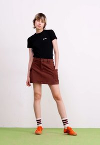 T-shirt nera con logo, gonna marrone con tasche, scarpe arancioni, calze a righe. Modella in piedi contro uno sfondo bianco su pavimento verde.