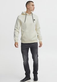 Felpa con cappuccio color crema con tasca frontale e lacci neri, abbinata a jeans scuri e sneaker nere. Design semplice e informale.