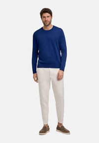 Navyblauer gestrickter Pullover mit Rundhalsausschnitt, kombiniert mit cremefarbenen, schmal zulaufenden Hosen und braunen Slippern, vor einem schlichten Hintergrund stehend.