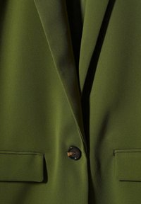 Gros plan d'un blazer vert olive avec un bouton noir unique et deux poches à rabat de chaque côté.