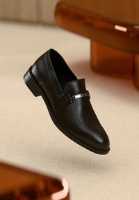 Svart skinnloafer med detalj av sølvfarget metallbit svevende over en beige overflate, med uskarp brun møbel i bakgrunnen.