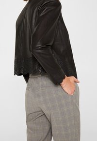 Chaqueta de cuero negra con dobladillo scalloped y bordado. El diseño presenta una silueta ajustada y una textura suave, mostrada con pantalones grises de estampado.