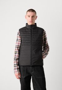 Colete puffer preto com gola alta, fecho de correr e textura acolchoada. Usado sobre uma camisa de padrão xadrez multicolorido e combinado com jeans escuros.