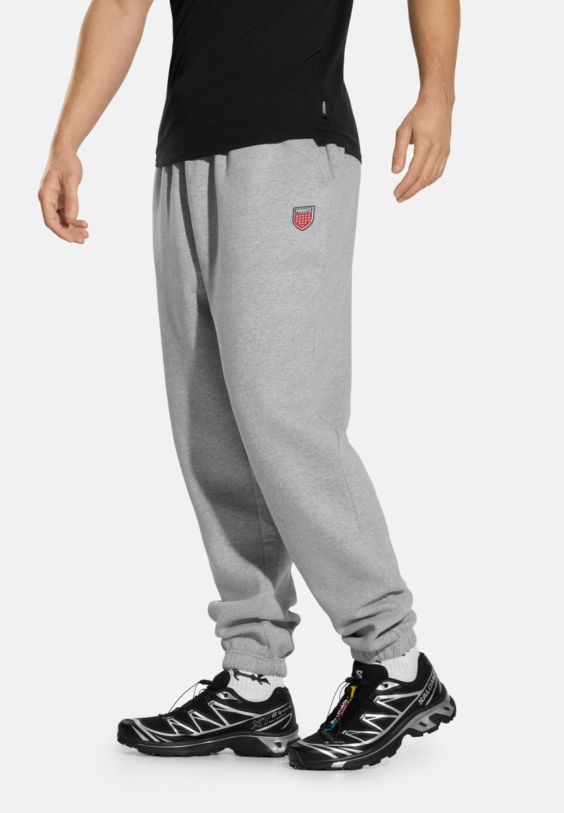 Szare spodnie dresowe z elastycznym pasem, ozdobione małym czerwonym emblematem. Do tego czarne buty sportowe z odblaskowymi akcentami i sznurowadłami.