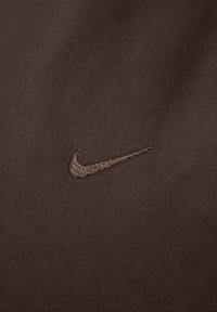 Hnědá tkanina s diagonálním tkaním má vyšitý logo Nike ve světlejším hnědém odstínu, umístěné v pravém dolním rohu.