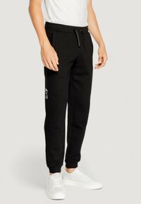 Black sweatpants with a tapered fit, drawstring waist, side pockets, and white logo accents on the leg, paired with white sneakers.