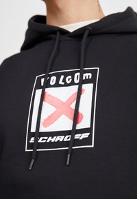 Sudadera negra con un logo gráfico que contiene una "X" roja y el texto "VOLCOM SCHROEF". El diseño incluye cordones y una textura suave.