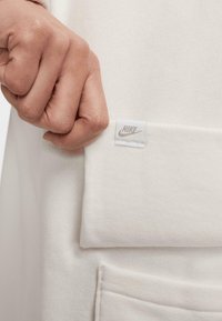 Fehér pulóver, texturált felülettel, látható Nike címkével. Az anyagot a kéz a szegélyénél fogja, bemutatva annak puha anyagát.
