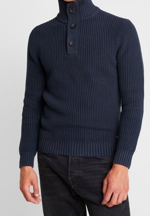 Navyblauw gebreid sweater met een hoge kraag, geribbelde manchetten en drie knopen aan de hals. Geweven patroon over het gehele oppervlak.