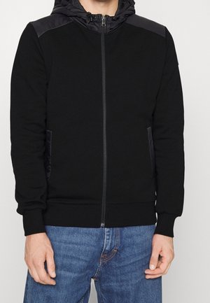 Mann trägt eine schwarze Kapuzenjacke mit Reißverschluss und gepolsterten Schultern sowie blaue Jeans, stehend vor einem schlichten Hintergrund.