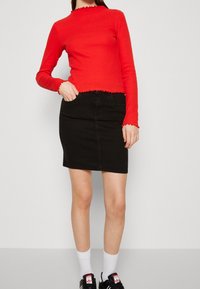 Czerwony sweter z długim rękawem i falbaniastym dekoltem, w połączeniu z czarną spódnicą dżinsową. Modelka nosi białe skarpetki i czarne trampki.