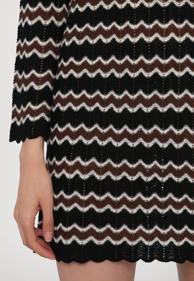 Robe tricotée à motif zigzag noir, marron et blanc avec un ourlet en forme de vague, présentant un design texturé à mailles ouvertes.