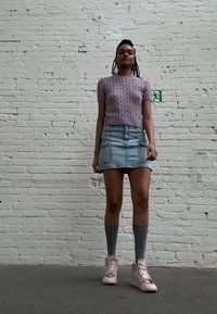 Ljuslila stickad kortärmad topp, denimminikjol, grå knälånga strumpor, rosa högskoor, ståendes mot en vit tegelvägg.