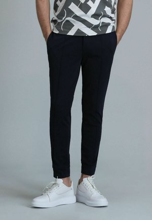 Pantalons bleu marine fuselés avec poches latérales, en tissu structuré et détails de plis, associés à des baskets blanches avec un design texturé.