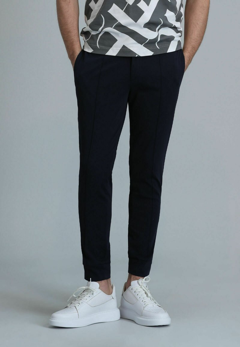 Marineblaue, schmal zulaufende Hosen mit Seitentaschen, strukturierter Stoff und Falten-Details, kombiniert mit weißen Sneakers mit strukturiertem Design.