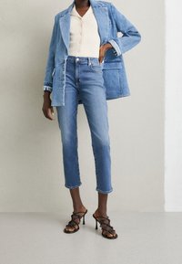 Džínová bunda ve světle modré barvě se strukturovaným designem, spárovaná s tmavě modrými zkrácenými džíny. Celkový vzhled doplňují černé sandály na vysokém podpatku se strapci.