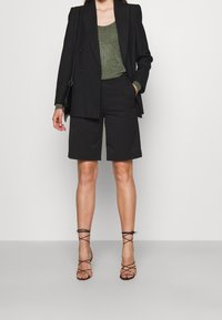 Blazer noir sur mesure porté avec un haut vert métallique, associé à un short noir et des talons noirs à lanières. Tissu lisse et design structuré.