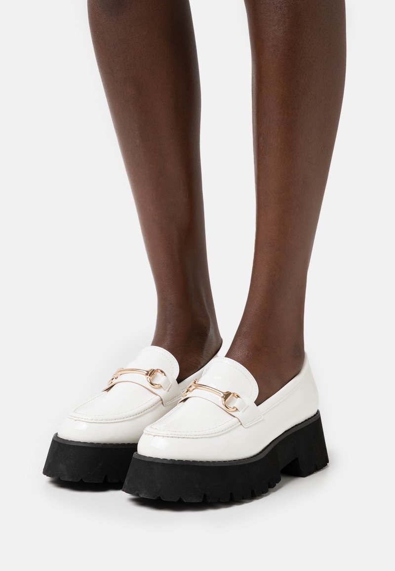 Jambes à la peau foncée portant des mocassins à plateforme en cuir blanc avec des détails en métal doré et des semelles épaisses noires sur un fond clair.
