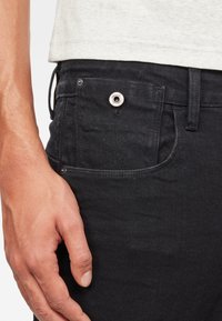 Jeans de mezclilla negra con un bolsillo frontal con un remache de metal plateado, textura suave y un lazo para cinturón, mostrados con una mano descansando cerca.