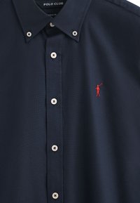 Marineblaue Hemdjacke aus Baumwolle mit strukturiertem Finish, ausgestattet mit weißen Knöpfen und einem kleinen roten Logo, das auf der Brust gestickt ist.