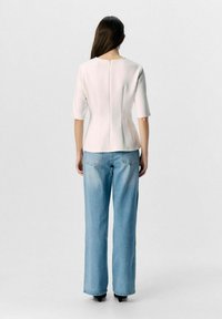 Weißes, tailliertes Oberteil mit kurzen Ärmeln, Reißverschluss im Rücken und Nahtdetails, kombiniert mit hellblauen, weit geschnittenen Jeans. Glattes Material, minimalistisches Design.