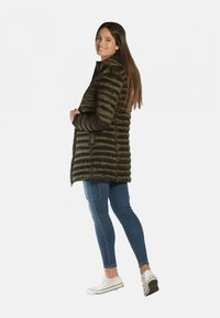 Veste rembourrée vert foncé avec un design à carreaux horizontaux. Associée à un jean bleu et des baskets blanches, elle se distingue par un col montant et des zips.