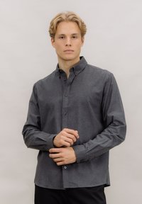 Grå skjorte med knapper laget av teksturert stoff, med spredd krage, lange ermer og rett kant. Knappelukkinger foran.