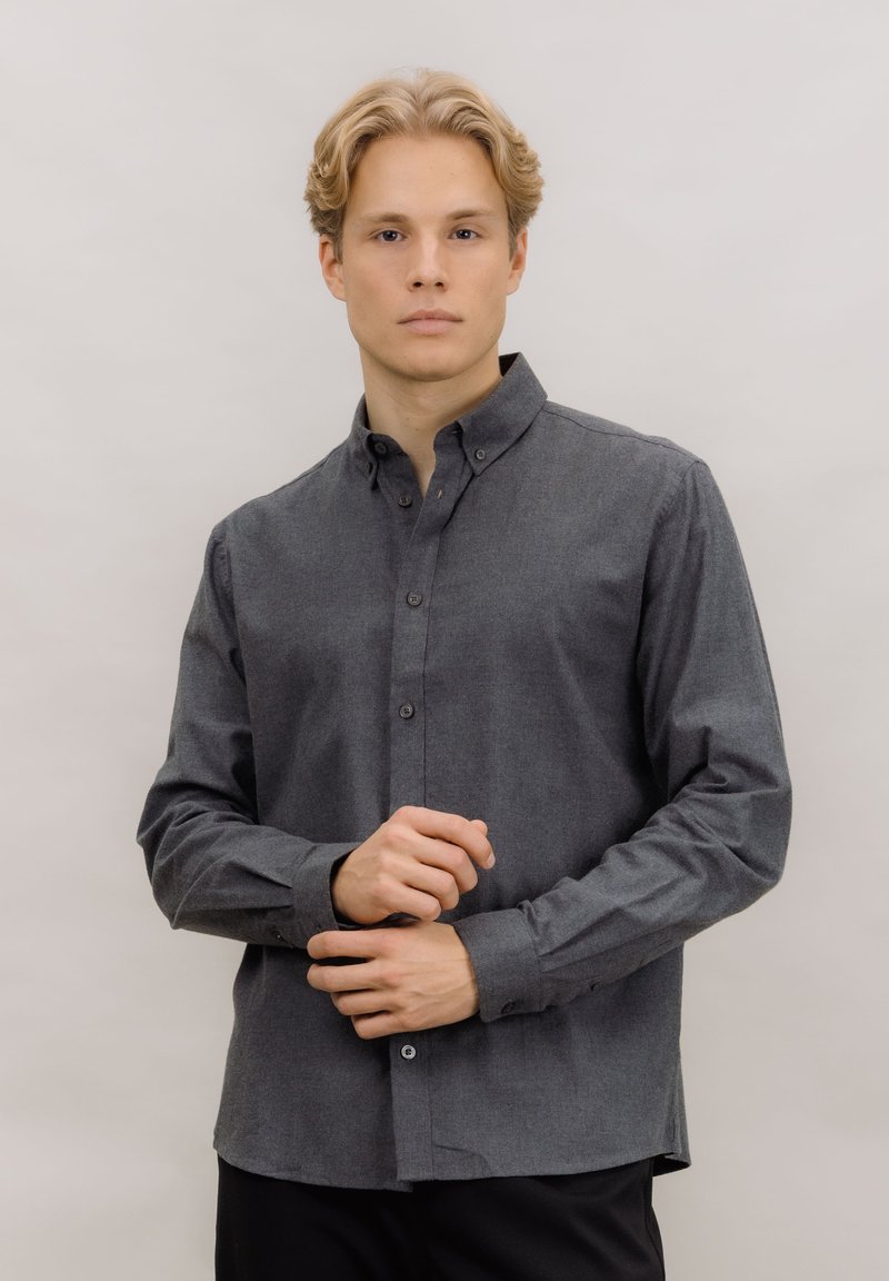 Grå skjorte med knapper laget av teksturert stoff, med spredd krage, lange ermer og rett kant. Knappelukkinger foran.