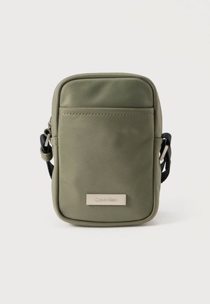 Sac bandoulière en nylon vert olive avec une texture lisse, doté d'une poche zippée à l'avant et d'une plaque logo Calvin Klein argentée. Sangle ajustable incluse.