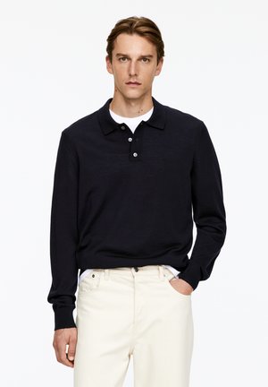 Marineblaue Langarm-Poloshirt aus einem weichen Stoff, mit einer Knopfleiste mit drei Knöpfen und gerippten Bündchen, kombiniert mit cremefarbenen Hosen.