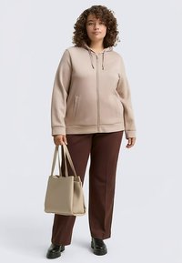Felpa con cappuccio beige con zip, polsini a coste e tasche frontali, abbinata a pantaloni marroni e una borsa a tracolla beige, indossata con scarpe nere.