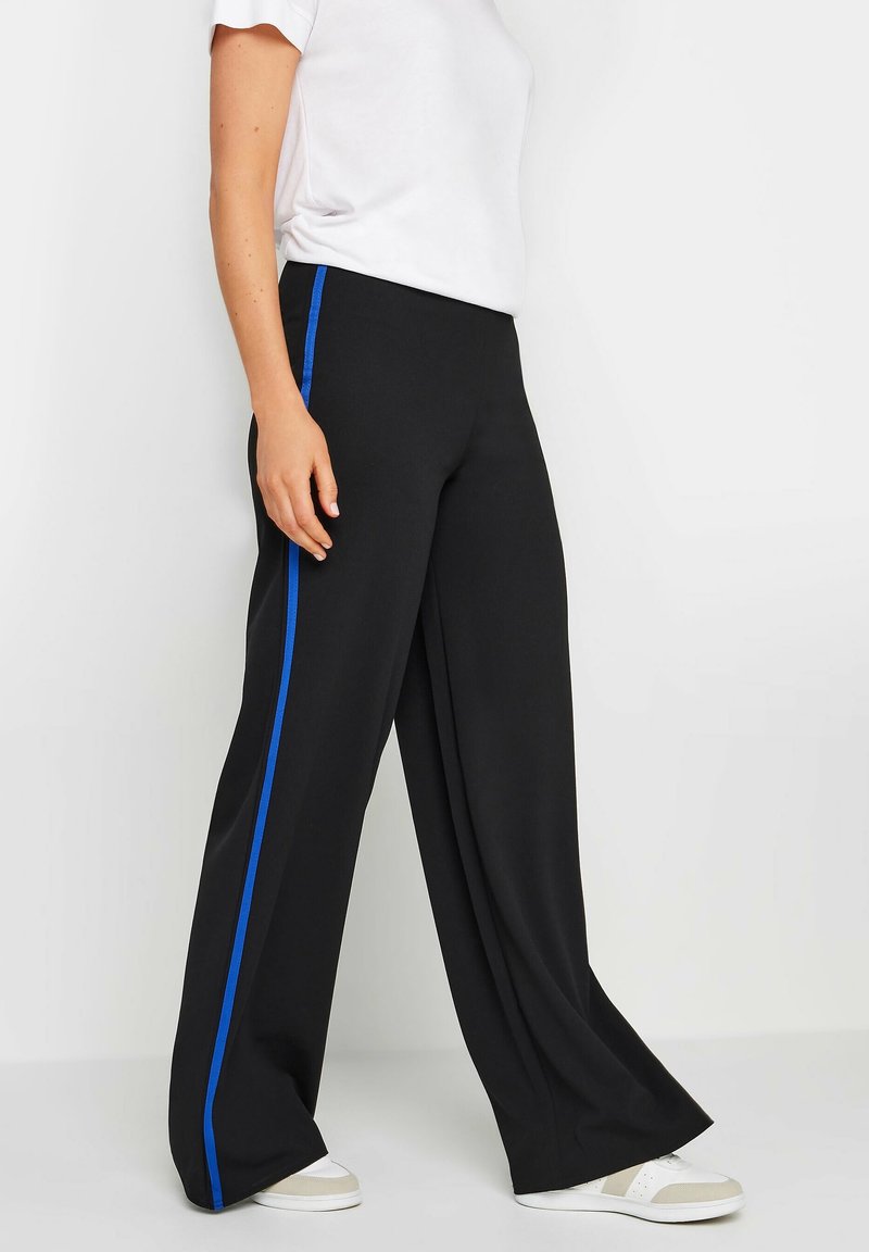 Personne portant un pantalon noir à jambes larges avec une bande latérale bleue, des baskets blanches et une chemise blanche à manches courtes, debout devant un fond blanc.