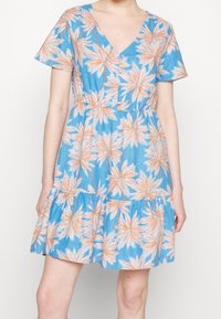 Femme portant une robe bleue à manches courtes avec un motif de feuilles de palmier florales pêche et des boutons sur la poitrine, sur fond uni.