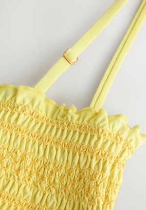 Sac en tissu smocké jaune avec des bords froncés, des sangles jaunes ajustables et des détails en matériel doré. Surface texturée avec un gathering élastique.