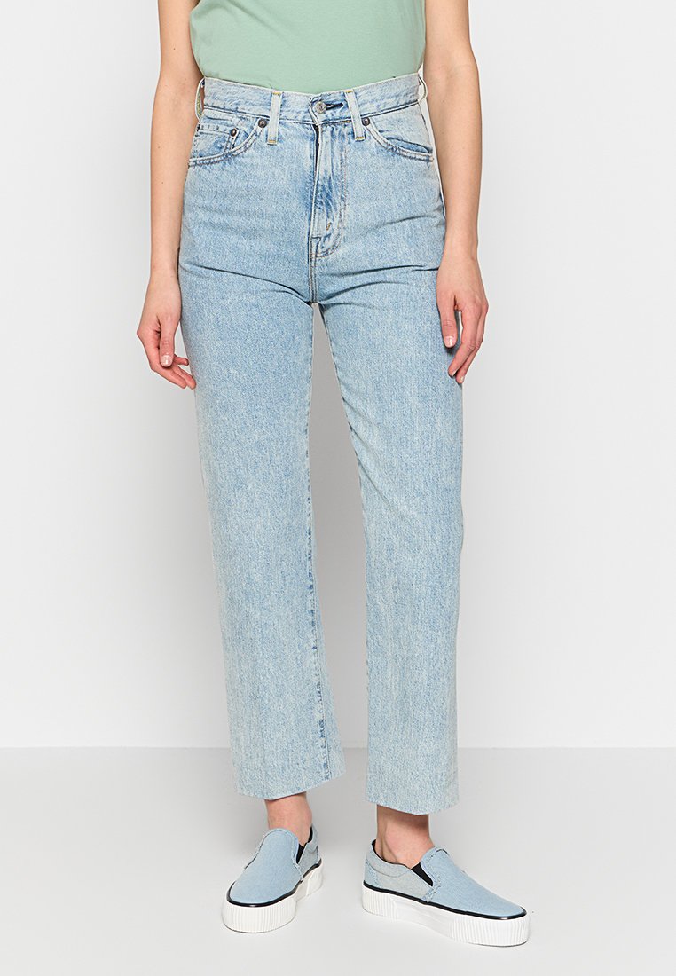 Jean taille haute bleu clair avec une coupe droite, présentant une fermeture zippée à l'avant et un style à cinq poches. Tissu denim texturé.